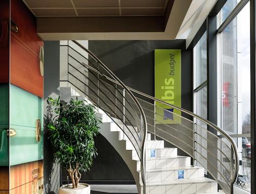 Bright stairwell with a metal railing and green “ibis budget” sign next to large windows.