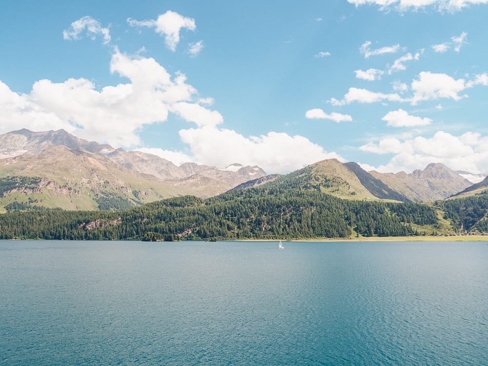 A peaceful mountain lake with clear blue water and a sailboat in the distance, surrounded by green forested hills and peaks under a bright, partly cloudy sky.